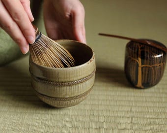 May include: A close-up of a hand holding a bamboo whisk over a green ceramic bowl with a dark liquid inside. The bowl is sitting on a woven mat. A small wooden container with a lid and a wooden spoon is in the background.