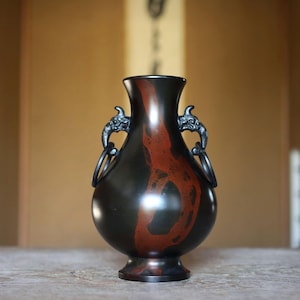 May include: A dark brown vase with a red pattern and two ornate handles. The vase has a bulbous body and a narrow neck. The handles are circular and attached to the vase with dragon-like figures. The vase is on a wooden surface.