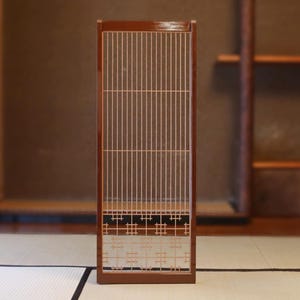 Puede incluir: Una pantalla Shoji japonesa rectangular y alta con un marco de madera marrón oscuro. La pantalla presenta listones verticales, delgados y de color claro, y un panel inferior decorativo con patrones geométricos. La pantalla está colocada en una habitación japonesa tradicional.