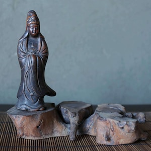 Pode incluir: Uma estátua de madeira esculpida de uma figura vestida com uma expressão serena, em pé sobre uma base de madeira rústica. A estátua é de cor castanho escuro e a base tem um aspeto natural e envelhecido. O fundo é um verde-azulado suave.