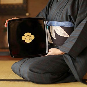 May include: A black lacquered tray with a gold floral emblem is held by a person wearing a dark blue kimono. The tray is square with rounded corners. The kimono has a striped pattern and a patterned obi.