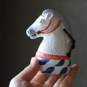 May include: A white ceramic horse figurine with a red and blue design. The horse has a gray mane and tail, and a red bridle. The figurine has Japanese text on its side and is held in a hand.