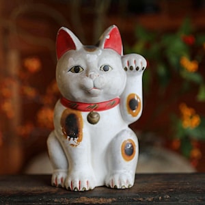 May include: A white ceramic Maneki Neko cat figurine with a raised paw. The cat has red accents on its ears and collar, with black and orange markings. A small gold bell hangs from the collar. The figurine is a symbol of good luck and fortune.