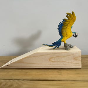 Pode incluir: Um calço de porta de madeira com acabamento natural, com uma figura colorida de arara em cima. A arara tem plumagem amarela e azul e um bico preto, em pose de voo. O calço de porta está sobre uma superfície de madeira.
