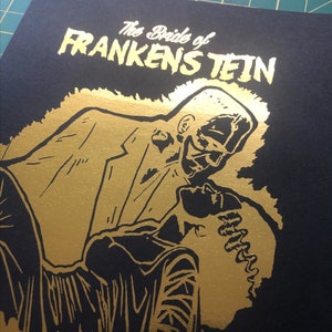 K&ouml;nnte beinhalten: Ein schwarzer und goldener Druck eines tanzenden Paares. Der Druck zeigt den Text "The Bride of Frankenstein" in goldenen Buchstaben.