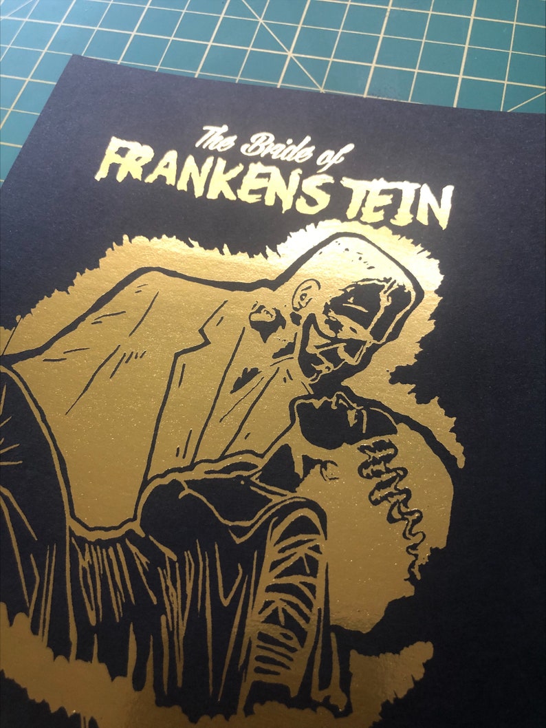 K&ouml;nnte beinhalten: Ein schwarzer und goldener Druck eines tanzenden Paares. Der Druck zeigt den Text "The Bride of Frankenstein" in goldenen Buchstaben.
