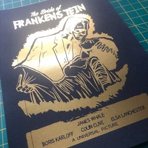 K&ouml;nnte beinhalten: Ein Goldfoliendruck des Filmplakats f&uuml;r "Die Braut des Frankenstein". Das Plakat zeigt ein Schwarzwei&szlig;bild des Monsters, das die Braut tr&auml;gt. Der Text auf dem Plakat lautet "Die Braut des Frankenstein", "James Whale", "Ein Universal-Film", "Colin Clive", "Elsa Lanchester" und "Boris Karloff".