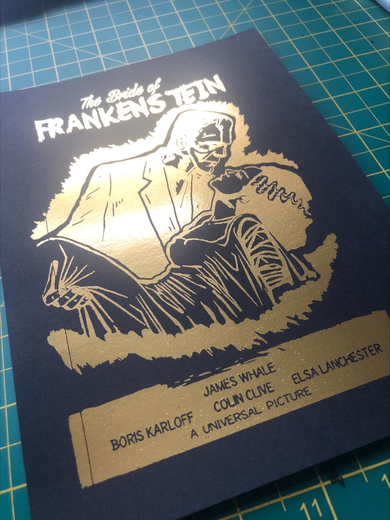 K&ouml;nnte beinhalten: Ein Goldfoliendruck des Filmplakats f&uuml;r "Die Braut des Frankenstein". Das Plakat zeigt ein Schwarzwei&szlig;bild des Monsters, das die Braut tr&auml;gt. Der Text auf dem Plakat lautet "Die Braut des Frankenstein", "James Whale", "Ein Universal-Film", "Colin Clive", "Elsa Lanchester" und "Boris Karloff".