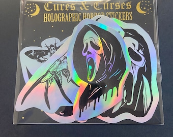 May include: A set of five holographic horror stickers featuring a screaming ghost, a moth, and a skull. The stickers are packaged in a clear plastic bag with a black background and the text "Cures & Curses Holographic Horror Stickers".