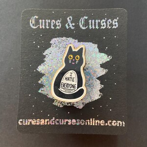 May include: A black cat enamel pin with the text "I HATE EVERYONE" on a white banner. The pin is on a black card with the words "Cures & Curses" and "curesandcursesonline.com" in silver lettering.