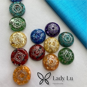 May include: A selection of colourful, round chakra stones with silver symbols. The stones are in shades of turquoise, green, yellow, red, orange, blue, and purple. The Lady Lu Handmade logo is in the bottom right corner.