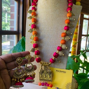 May include: A pink and orange silk thread necklace with gold accents and a matching pair of earrings. The necklace has a large gold pendant with intricate designs. The earrings have a similar design and feature a pink tassel. The necklace and earrings are displayed on a white mannequin and a yellow card with the text "Riaaj Boutique" and "Design. Treasure. Create."