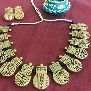 Op de afbeelding: Een gouden ketting met zwarte kralen en ovale hangers met geometrische patronen. De ketting ligt op een rode stoffen achtergrond.