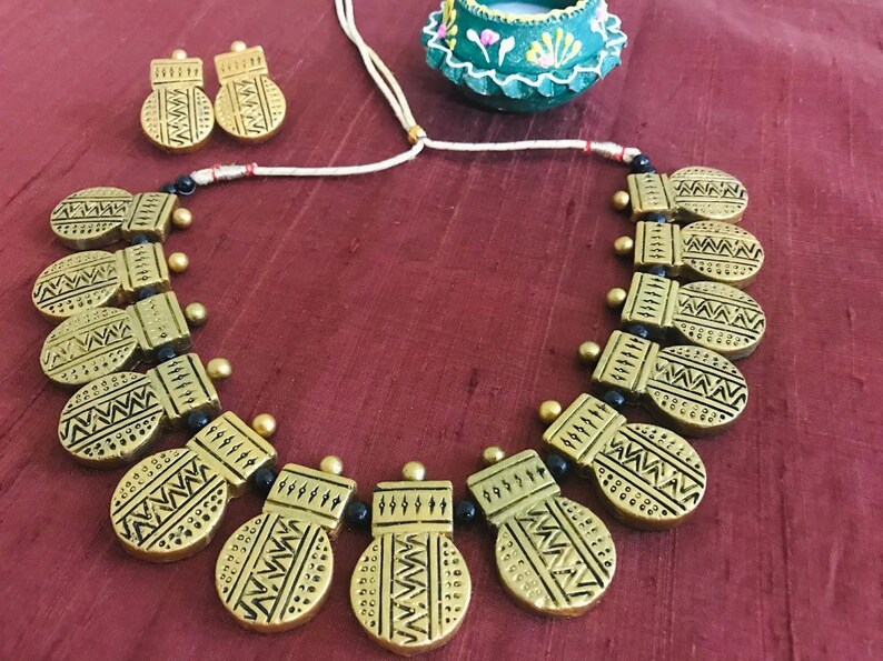 Op de afbeelding: Een gouden ketting met zwarte kralen en ovale hangers met geometrische patronen. De ketting ligt op een rode stoffen achtergrond.