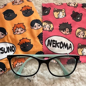 Może przedstawiać: Dwa mikrofibrowe ściereczki do czyszczenia z nadrukowanymi postaciami anime. Pomarańczowa ściereczka ma napis "Karasuno", a różowa "Nekoma". Na ściereczkach leży para czarnych i niebieskich okularów.