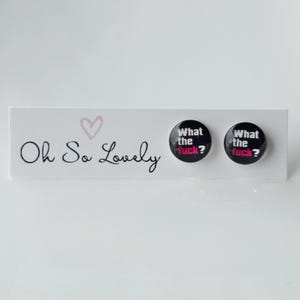 May include: Two black round buttons with the text "What the Fuck?" in white and pink, presented on a white card with the words "Oh So Lovely" and a pink heart. These are likely accessories or pins.