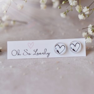 May include: A pair of clear stud earrings with a black heart and arrow design. The earrings are displayed on a white card with the text "Oh So Lovely" and a pink heart. The background is a soft, neutral color with white flowers.