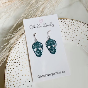 May include: Teal blue skull earrings with a delicate floral design. The earrings are on a white card with the text "Oh So Lovely" and the website "Ohsolovelyonline.ca".