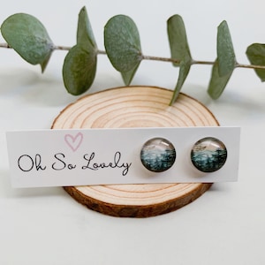 May include: A pair of stud earrings featuring a forest scene image. The earrings are displayed on a white card with the text "Oh So Lovely" in black.