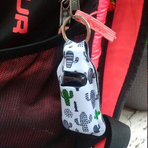 May include: A white and black cactus print fabric keychain with a black and silver hand sanitizer bottle inside. The keychain is attached to a black backpack with a pink zipper.