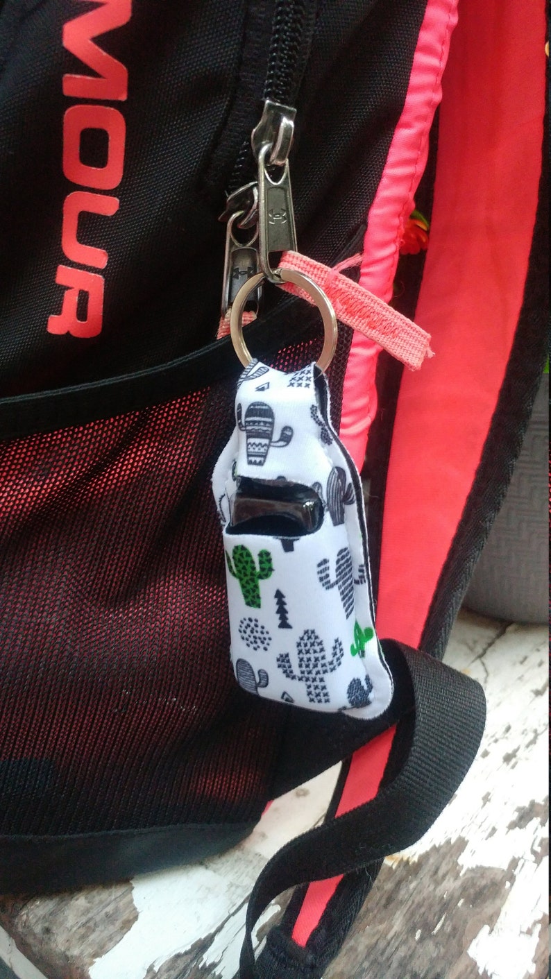 May include: A white and black cactus print fabric keychain with a black and silver hand sanitizer bottle inside. The keychain is attached to a black backpack with a pink zipper.