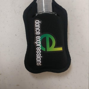 May include: Black hand sanitizer holder with a key ring and a clear plastic bottle. The holder has the text "dance expressions" and a green and yellow logo.