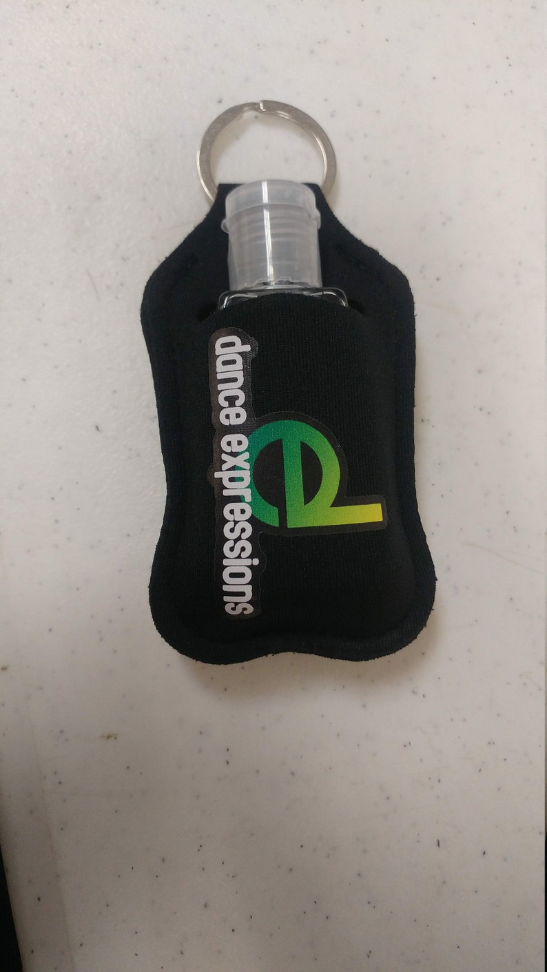 May include: Black hand sanitizer holder with a key ring and a clear plastic bottle. The holder has the text "dance expressions" and a green and yellow logo.