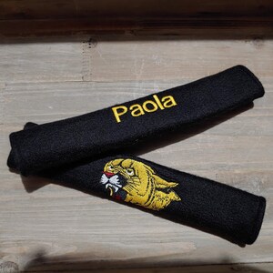 Puede incluir: Dos fundas de cinturón de seguridad de coche de tela negra. Una funda tiene el nombre "Paola" bordado en hilo amarillo. La otra funda tiene una imagen bordada en amarillo de una cabeza de león.