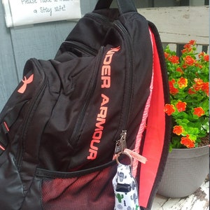May include: Black Under Armour backpack with a red zipper and a red mesh pocket. The backpack has a small white and green cactus-patterned keychain attached to it. The backpack is sitting on a wooden surface.