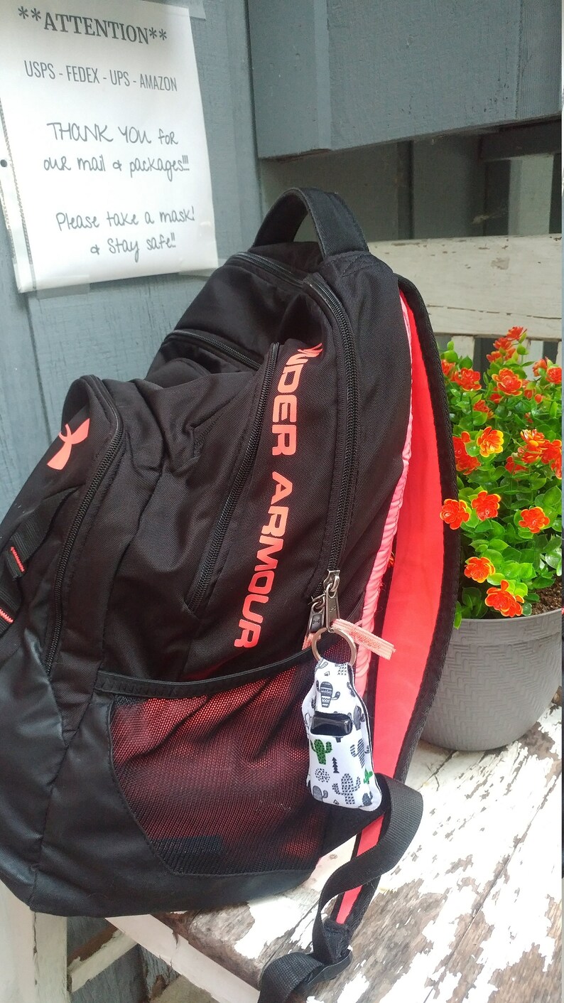 May include: Black Under Armour backpack with a red zipper and a red mesh pocket. The backpack has a small white and green cactus-patterned keychain attached to it. The backpack is sitting on a wooden surface.