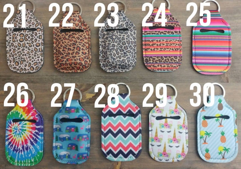 May include: Ten colorful hand sanitizer holders with keychains. The holders are made of fabric and have various patterns, including animal print, tie-dye, and geometric designs. Each holder is numbered from 21 to 30.