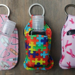 May include: Three hand sanitizer holders with keychains. The holders are made of fabric and have different patterns: medical symbols, puzzle pieces, and pink ribbons. Each holder has a clear plastic bottle of hand sanitizer inside.