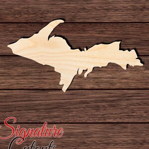 May include: A wooden cutout of the Upper Peninsula of Michigan. The cutout is a light brown color and is on a dark brown wood background. The text "Signature Cutouts" is visible in the bottom left corner of the image.