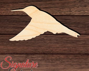 May include: A flat, light-colored wooden cutout in the shape of a hummingbird in flight. The silhouette features a long, thin beak, a rounded head, a large wing extended downward, and a pointed tail. The surface shows a natural wood grain pattern.