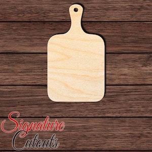 May include: A light-colored, unfinished wooden cutout in the shape of a rectangular cutting board with rounded corners and a handle at the top featuring a circular hole for hanging. The wood grain is visible across the flat surface of the object.