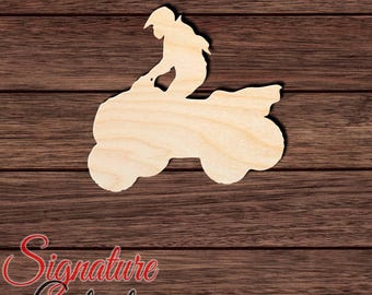 May include: A light-colored, flat wooden cutout in the shape of a person riding an all-terrain vehicle. The silhouette shows the rider leaning forward on the four-wheeled vehicle, with visible wood grain texture throughout the surface of the cutout.