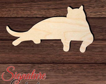 May include: A light-colored, flat wooden cutout in the shape of a cat lying down in a relaxed position. The silhouette shows the cat with its tail extended, ears pointed upward, and legs tucked underneath its body, displaying a natural wood grain.