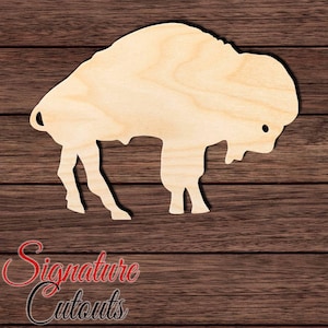 May include: A light-colored, flat wooden cutout in the shape of a buffalo or bison. The silhouette features the distinct hump, head, and four legs of the animal, with a small circular hole near the head area. The wood grain is visible throughout.