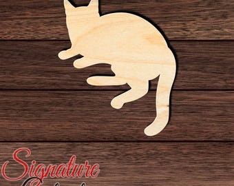 May include: A light-colored, flat wooden cutout in the shape of a cat lying down. The cat is depicted from a top-down perspective with its head turned, ears pointed, and tail extended downwards. The wood grain is visible across the surface.