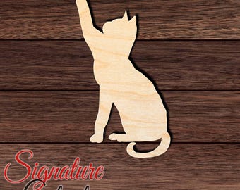 May include: A light-colored wooden cutout in the shape of a cat sitting with one front paw raised upward. The cat silhouette features pointed ears, a curved tail, and a smooth, natural wood grain finish, presented as a flat decorative object.
