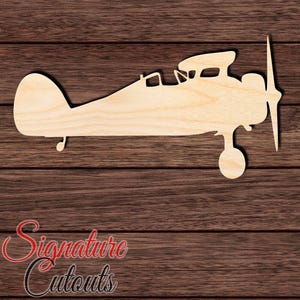 May include: A light-colored wooden cutout in the shape of a vintage airplane, featuring a side profile view with a single propeller, a cockpit opening, a rounded tail section, and a single visible landing gear wheel positioned beneath the fuselage.