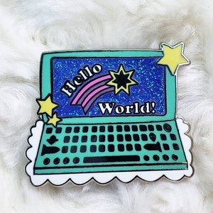 Könnte beinhalten: Eine grüne und blaue Emaille-Anstecknadel in Form eines Laptops mit einem weißen Wolkenrand. Der Bildschirm zeigt den Text "Hello World!" mit einem Regenbogen und einem Stern.