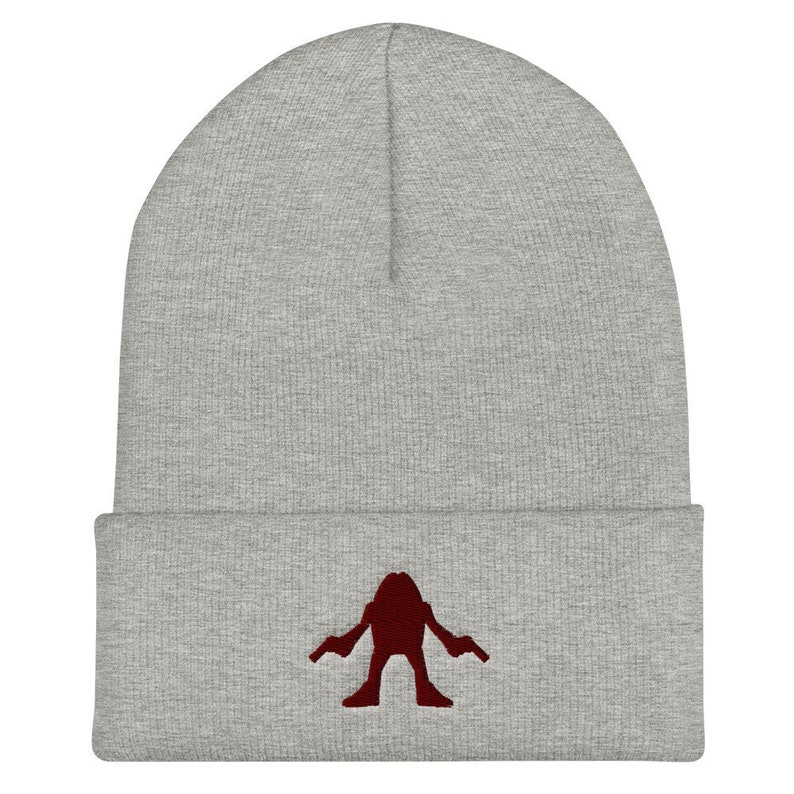 May include: Gray beanie with a red embroidered silhouette of a figure holding two guns.