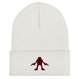 Könnte beinhalten: Weiße Strickmütze mit einer roten gestickten Silhouette einer Kreatur, die zwei Pistolen hält.
