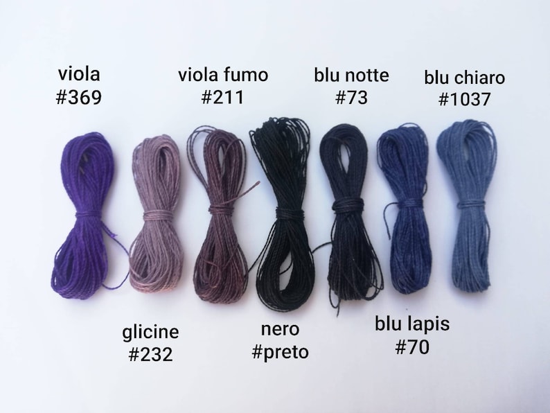 K&ouml;nnte beinhalten: Eine Sammlung von acht Spulen Stickgarn in verschiedenen Schattierungen von Lila, Blau und Schwarz. Die Spulen sind mit italienischen Namen und entsprechenden Farbcodes beschriftet.