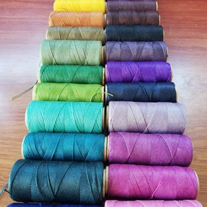 K&ouml;nnte beinhalten: Eine Sammlung von Spulen mit gewachstem Garn in verschiedenen Farben, darunter Gr&uuml;n, Blau, Lila, Rosa und Braun. Die Spulen sind in einer Reihe angeordnet, wobei die Farben allm&auml;hlich von hell nach dunkel &uuml;bergehen.