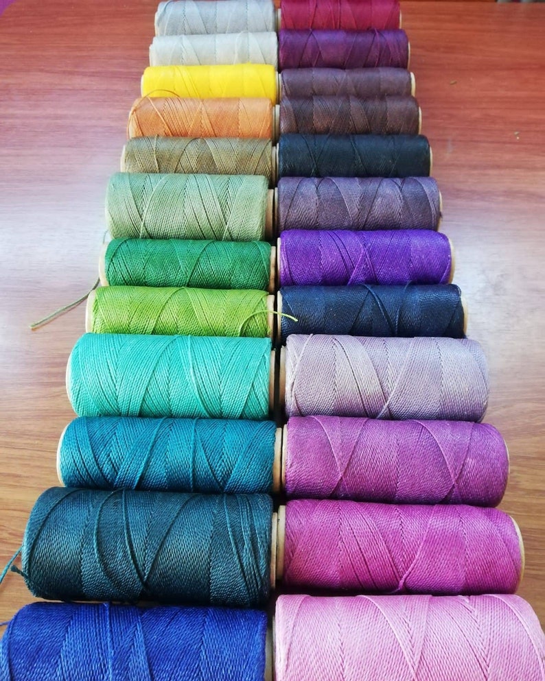 K&ouml;nnte beinhalten: Eine Sammlung von Spulen mit gewachstem Garn in verschiedenen Farben, darunter Gr&uuml;n, Blau, Lila, Rosa und Braun. Die Spulen sind in einer Reihe angeordnet, wobei die Farben allm&auml;hlich von hell nach dunkel &uuml;bergehen.