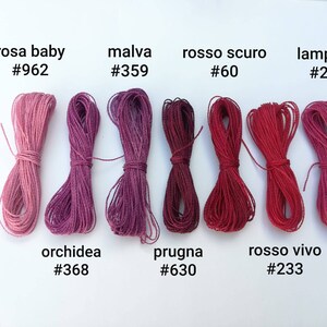 K&ouml;nnte beinhalten: Eine Sammlung von acht Spulen Stickgarn in Rosat&ouml;nen, Rot und Lila. Das Garn ist mit italienischen Namen und entsprechenden Farbcodes gekennzeichnet.