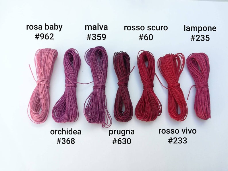 K&ouml;nnte beinhalten: Eine Sammlung von acht Spulen Stickgarn in Rosat&ouml;nen, Rot und Lila. Das Garn ist mit italienischen Namen und entsprechenden Farbcodes gekennzeichnet.