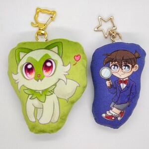 May include: Two plush keychains. One is green and features a cartoon white fox with red eyes. The other is blue and features a cartoon boy with brown hair wearing a blue jacket and holding a magnifying glass.